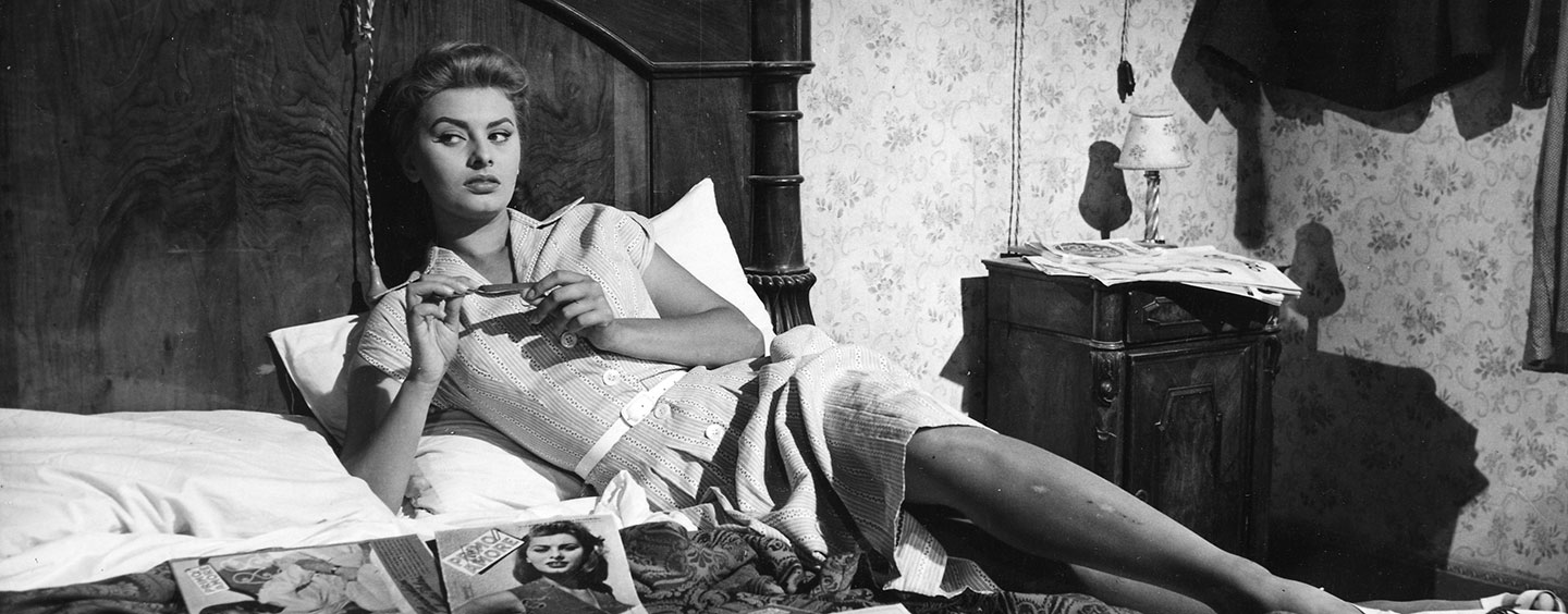 Black and white film frame of a woman laying in a bed with magazines strewn across it